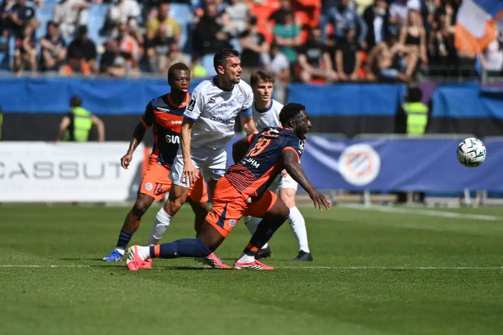 Soccer players from two teams chase the ball during a daytime match on a green field with a cheering crowd in the stands behind them.