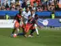 Soccer players from two teams chase the ball during a daytime match on a green field with a cheering crowd in the stands behind them.