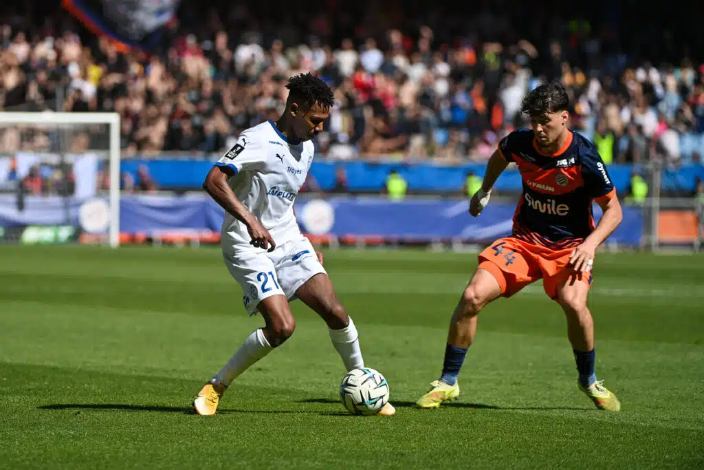 Two footballers duel for the ball on a sunny pitch, one in white with #21 and the other in navy and orange with 'smile' on the chest, crowd in the stands behind.