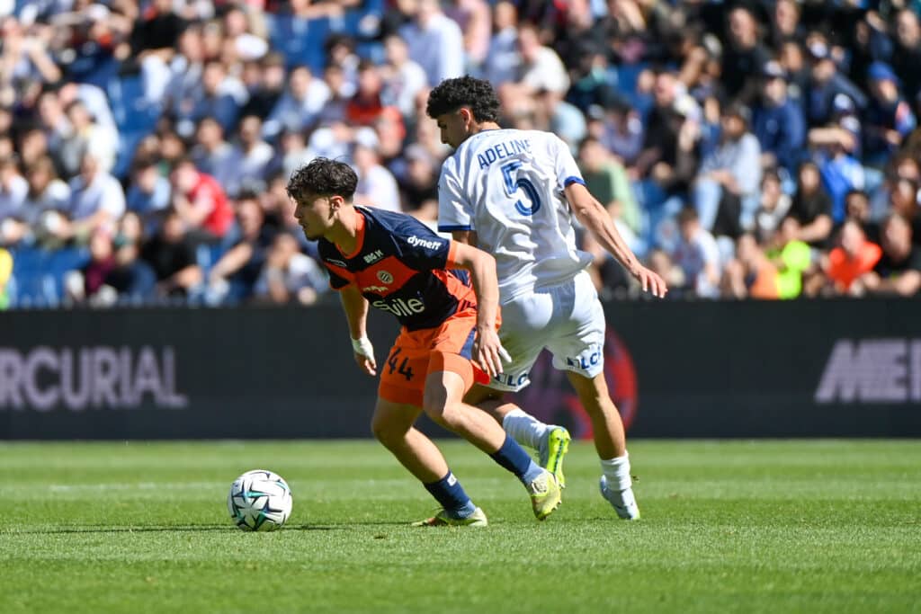 Chennahi Montpellier Herault SC v ESTAC Troyes - Ligue 2 BKT 20252026 | AllezPaillade.com Two soccer players compete for the ball on a sunny pitch, one in navy and orange kit and the other in a white kit with the number 5 and the name Adeline on the back, as a crowded stadium watches nearby and the ball rolls between them.