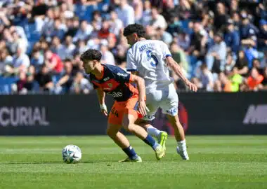 Two soccer players compete for the ball on a sunny pitch, one in navy and orange kit and the other in a white kit with the number 5 and the name Adeline on the back, as a crowded stadium watches nearby and the ball rolls between them.