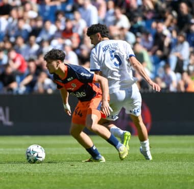 Two soccer players compete for the ball on a sunny pitch, one in navy and orange kit and the other in a white kit with the number 5 and the name Adeline on the back, as a crowded stadium watches nearby and the ball rolls between them.
