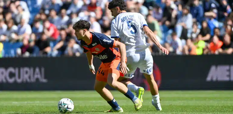 Two soccer players compete for the ball on a sunny pitch, one in navy and orange kit and the other in a white kit with the number 5 and the name Adeline on the back, as a crowded stadium watches nearby and the ball rolls between them.