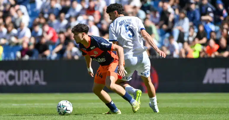 Two soccer players compete for the ball on a sunny pitch, one in navy and orange kit and the other in a white kit with the number 5 and the name Adeline on the back, as a crowded stadium watches nearby and the ball rolls between them.