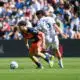 Two soccer players compete for the ball on a sunny pitch, one in navy and orange kit and the other in a white kit with the number 5 and the name Adeline on the back, as a crowded stadium watches nearby and the ball rolls between them.
