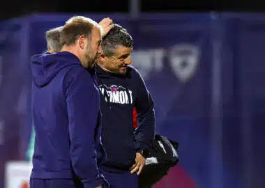 Two men in navy sportswear stand close together on a field, one patting the other's head in a casual coaching moment of discussion and camaraderie on the sidelines.