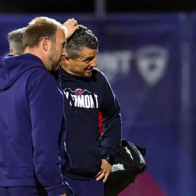 Two men in navy sportswear stand close together on a field, one patting the other's head in a casual coaching moment of discussion and camaraderie on the sidelines.