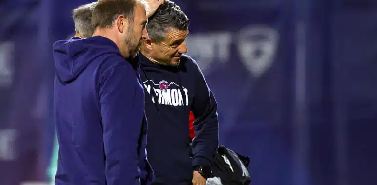 Two men in navy sportswear stand close together on a field, one patting the other's head in a casual coaching moment of discussion and camaraderie on the sidelines.