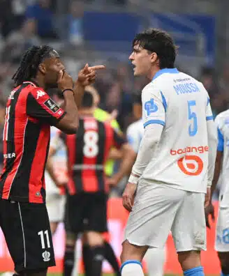 Two soccer players confront each other on the field, one in a red-and-black kit pointing at the other in a white-and-blue kit.