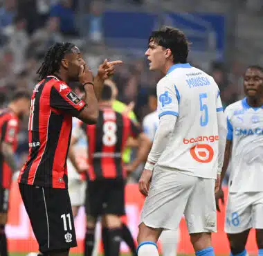 Two soccer players confront each other on the field, one in a red-and-black kit pointing at the other in a white-and-blue kit.