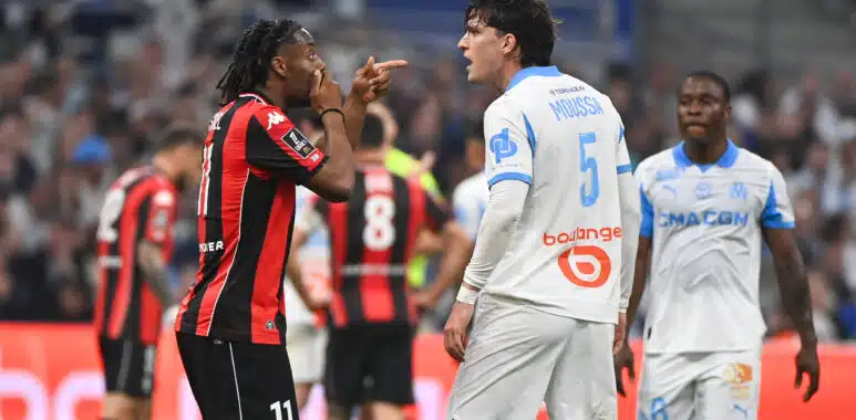 Two soccer players confront each other on the field, one in a red-and-black kit pointing at the other in a white-and-blue kit.