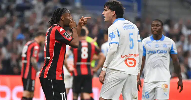 Two soccer players confront each other on the field, one in a red-and-black kit pointing at the other in a white-and-blue kit.