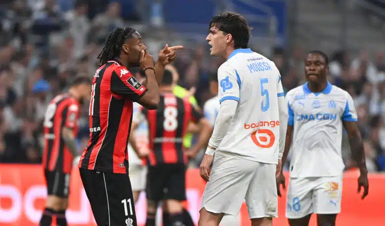 Two soccer players confront each other on the field, one in a red-and-black kit pointing at the other in a white-and-blue kit.