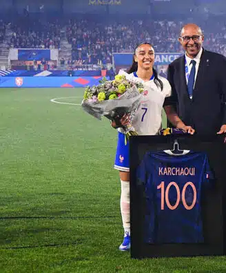 France football player in white jersey with bouquet of flowers, posing with a suited official beside a framed blue jersey reading 'KARCHAQUI 100' on a soccer field ceremony.