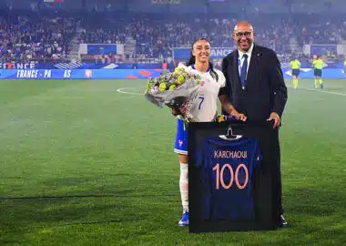 France football player in white jersey with bouquet of flowers, posing with a suited official beside a framed blue jersey reading 'KARCHAQUI 100' on a soccer field ceremony.