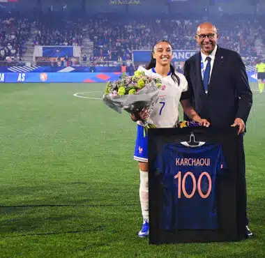 France football player in white jersey with bouquet of flowers, posing with a suited official beside a framed blue jersey reading 'KARCHAQUI 100' on a soccer field ceremony.