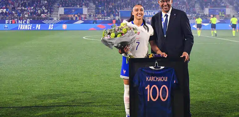 France football player in white jersey with bouquet of flowers, posing with a suited official beside a framed blue jersey reading 'KARCHAQUI 100' on a soccer field ceremony.