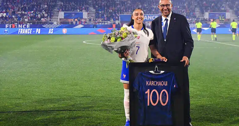 France football player in white jersey with bouquet of flowers, posing with a suited official beside a framed blue jersey reading 'KARCHAQUI 100' on a soccer field ceremony.