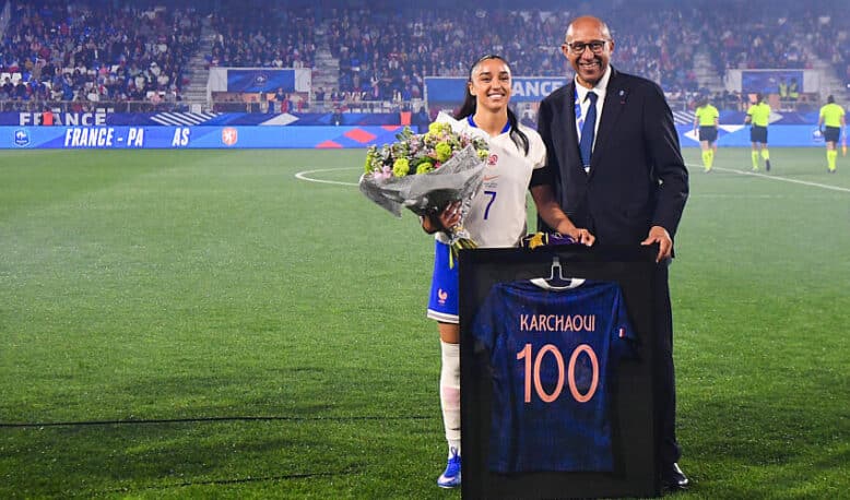 France football player in white jersey with bouquet of flowers, posing with a suited official beside a framed blue jersey reading 'KARCHAQUI 100' on a soccer field ceremony.