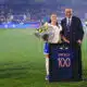 France football player in white jersey with bouquet of flowers, posing with a suited official beside a framed blue jersey reading 'KARCHAQUI 100' on a soccer field ceremony.
