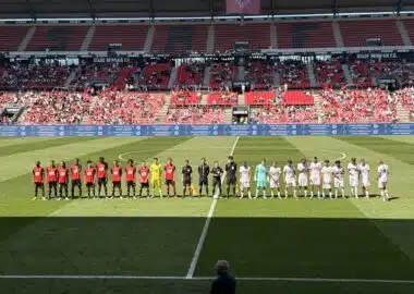 Football teams line up on the pitch before a match at a stadium, players in red and white kits facing forward with referees in between.
