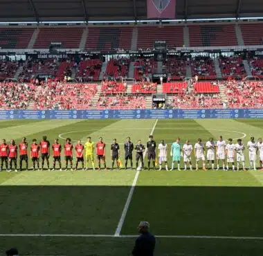 Football teams line up on the pitch before a match at a stadium, players in red and white kits facing forward with referees in between.
