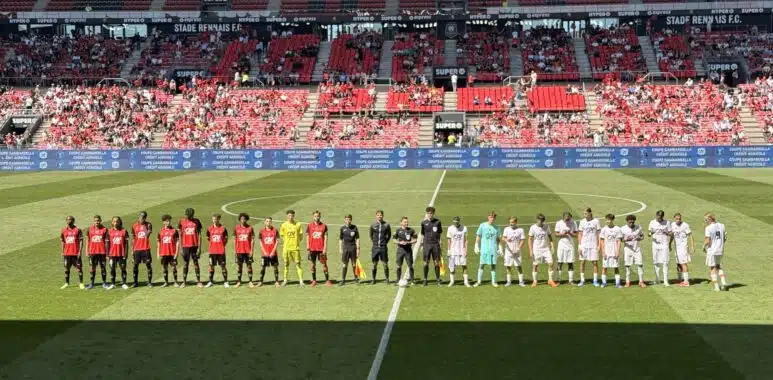 Football teams line up on the pitch before a match at a stadium, players in red and white kits facing forward with referees in between.