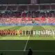 Football teams line up on the pitch before a match at a stadium, players in red and white kits facing forward with referees in between.
