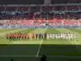 Football teams line up on the pitch before a match at a stadium, players in red and white kits facing forward with referees in between.