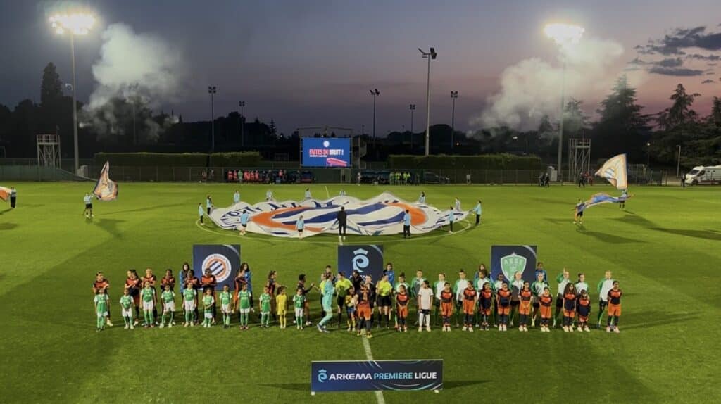Screenshot | AllezPaillade.com Teams in green and orange line up on a well-lit soccer pitch at dusk, with a large banner being opened behind them and banners in the foreground.