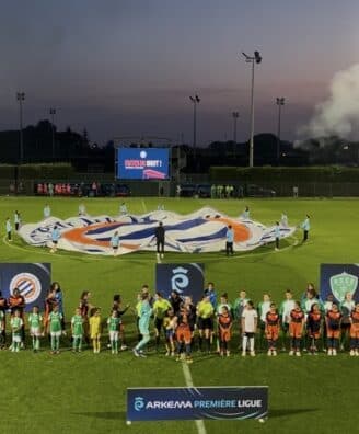Teams in green and orange line up on a well-lit soccer pitch at dusk, with a large banner being opened behind them and banners in the foreground.