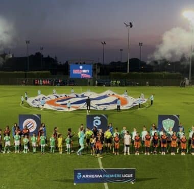 Teams in green and orange line up on a well-lit soccer pitch at dusk, with a large banner being opened behind them and banners in the foreground.