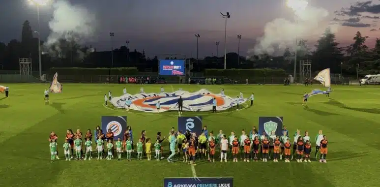 Teams in green and orange line up on a well-lit soccer pitch at dusk, with a large banner being opened behind them and banners in the foreground.