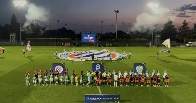 Teams in green and orange line up on a well-lit soccer pitch at dusk, with a large banner being opened behind them and banners in the foreground.