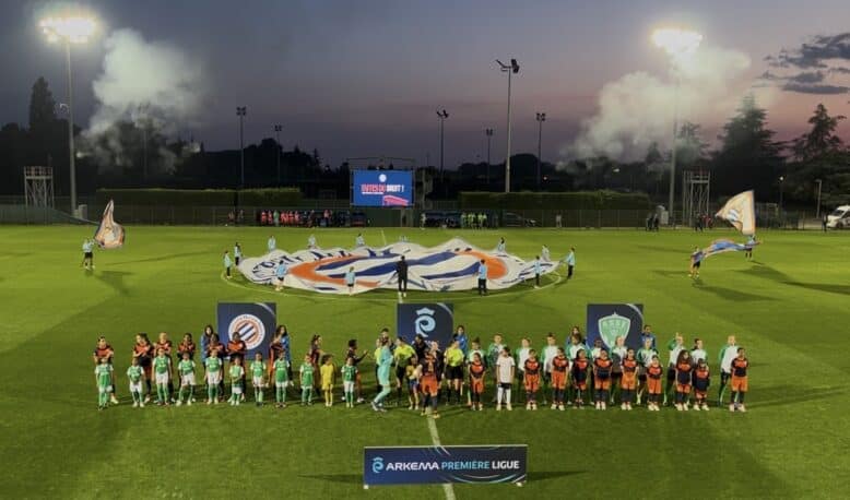 Teams in green and orange line up on a well-lit soccer pitch at dusk, with a large banner being opened behind them and banners in the foreground.