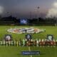 Teams in green and orange line up on a well-lit soccer pitch at dusk, with a large banner being opened behind them and banners in the foreground.