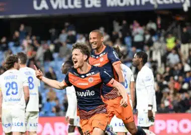 Two soccer players in orange and navy rush of celebration on the field as fans applaud in the stands, one pointing joyfully