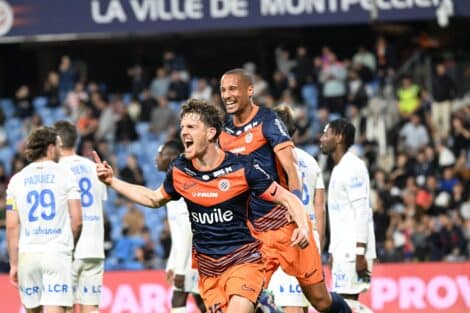 Two soccer players in orange and navy rush of celebration on the field as fans applaud in the stands, one pointing joyfully