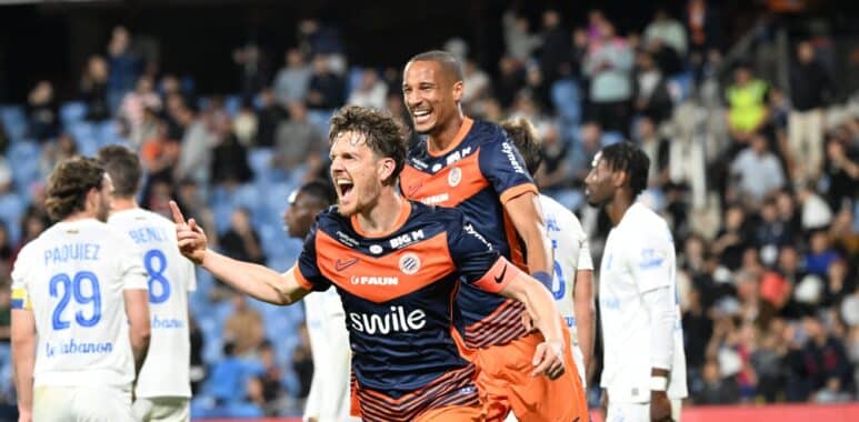 Two soccer players in orange and navy rush of celebration on the field as fans applaud in the stands, one pointing joyfully