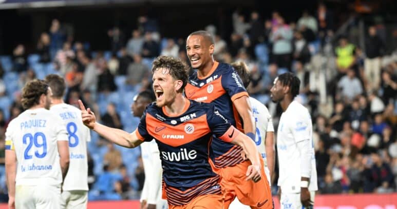 Two soccer players in orange and navy rush of celebration on the field as fans applaud in the stands, one pointing joyfully