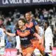 Two soccer players in orange and navy rush of celebration on the field as fans applaud in the stands, one pointing joyfully
