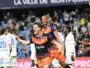 Two soccer players in orange and navy rush of celebration on the field as fans applaud in the stands, one pointing joyfully