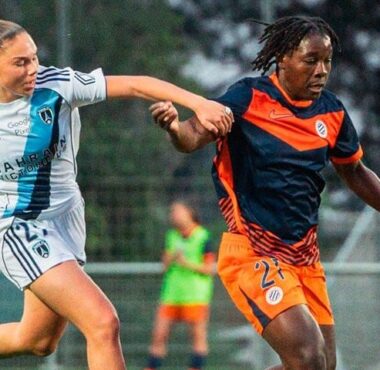 Two female soccer players race for the ball on a grass field during a match, one in white with blue stripes and the other in orange and navy.