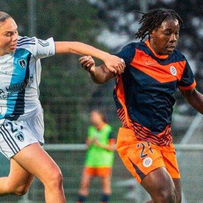 Two female soccer players race for the ball on a grass field during a match, one in white with blue stripes and the other in orange and navy.