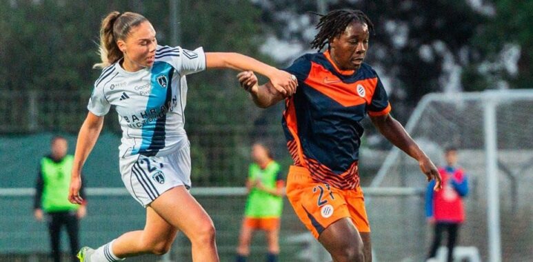 Two female soccer players race for the ball on a grass field during a match, one in white with blue stripes and the other in orange and navy.