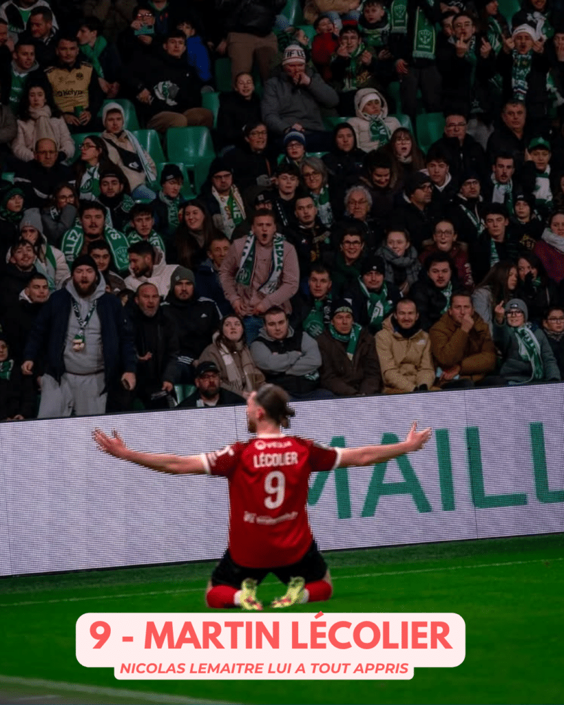 Soccer player in a red jersey number 9 kneels on the field with arms outstretched, facing a cheering crowd wearing green scarves, with a large banner displaying the name at the bottom.