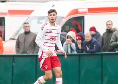 Soccer player in a white jersey with red shorts on the field, with spectators and an ambulance in the background behind a green barrier.