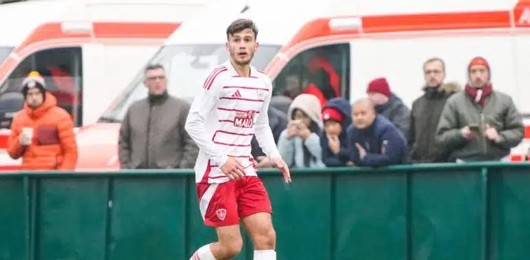 Soccer player in a white jersey with red shorts on the field, with spectators and an ambulance in the background behind a green barrier.