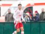 Soccer player in a white jersey with red shorts on the field, with spectators and an ambulance in the background behind a green barrier.