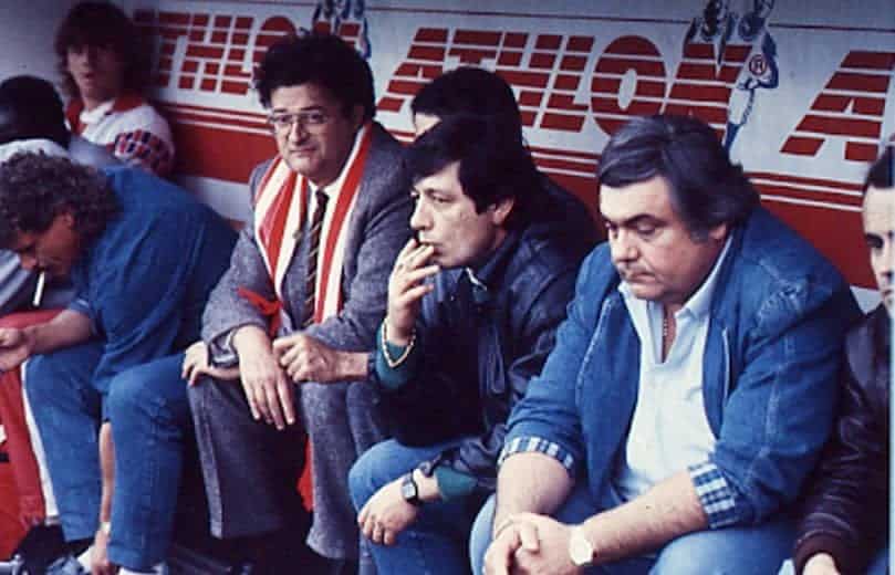 Men sitting on a sports bench/dugout, some smoking, with a red banner in the background.